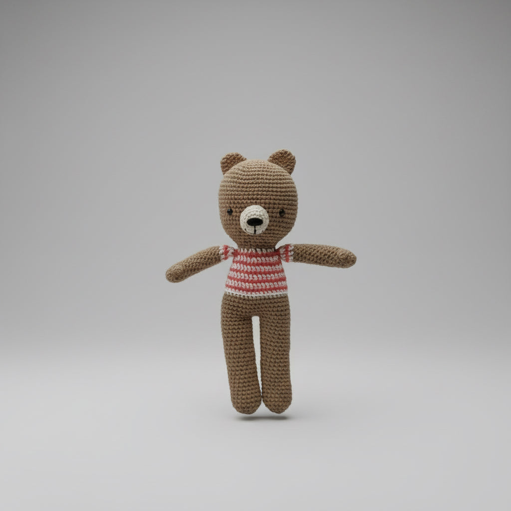 Hand holding a small crocheted teddy bear with a striped shirt against a plain background