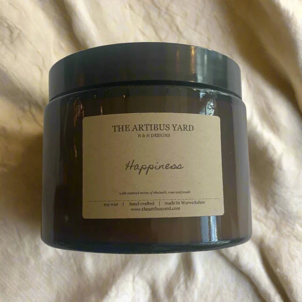 Hand-poured Happiness soy wax candle by The Artibus Yard, featuring notes of rhubarb, rose and musk; crafted in small batches in Warwickshire and poured into a reusable amber glass jar.